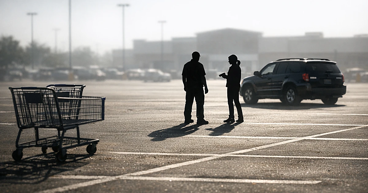 Two silhouetted figures standing apart in a sun-drenched parking lot with empty spaces and shopping carts nearby