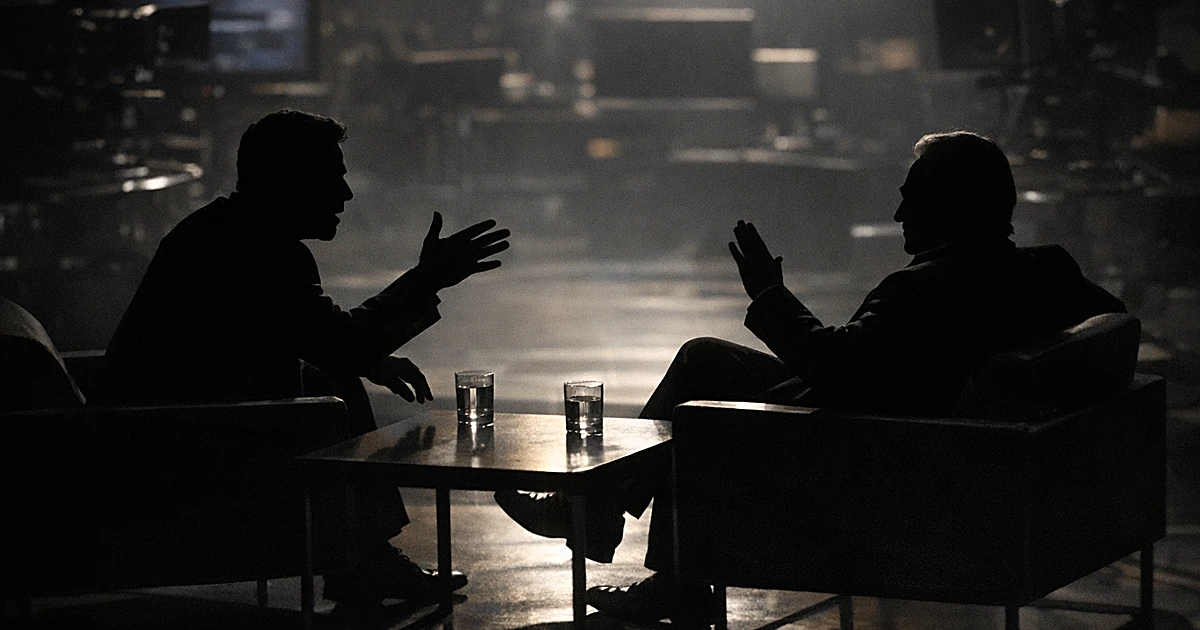 Two silhouetted figures in TV studio chairs during tense interview exchange with dramatic stage lighting