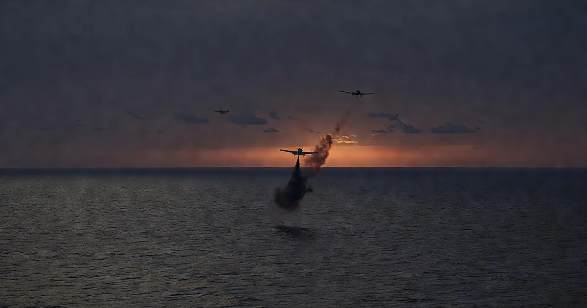 Aerial view of small aircraft trailing smoke over ocean waters at dusk with military jet silhouette in distance