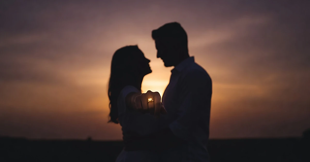 Silhouetted couple at sunset, woman's hand extended showing engagement ring catching light