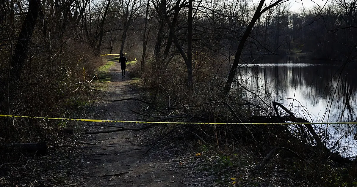 Wooded trail beside pond with police tape blocking path and distant runner silhouette among bare trees