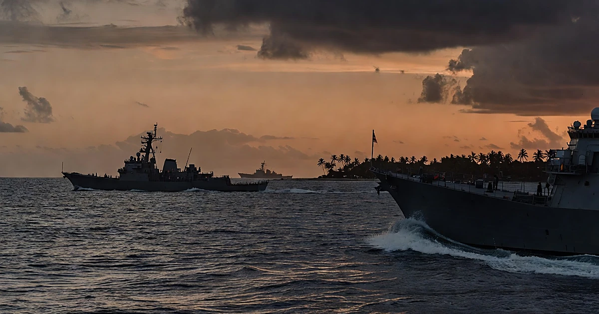 Military ships approaching a tropical island coastline at sunset with palm trees visible on shore